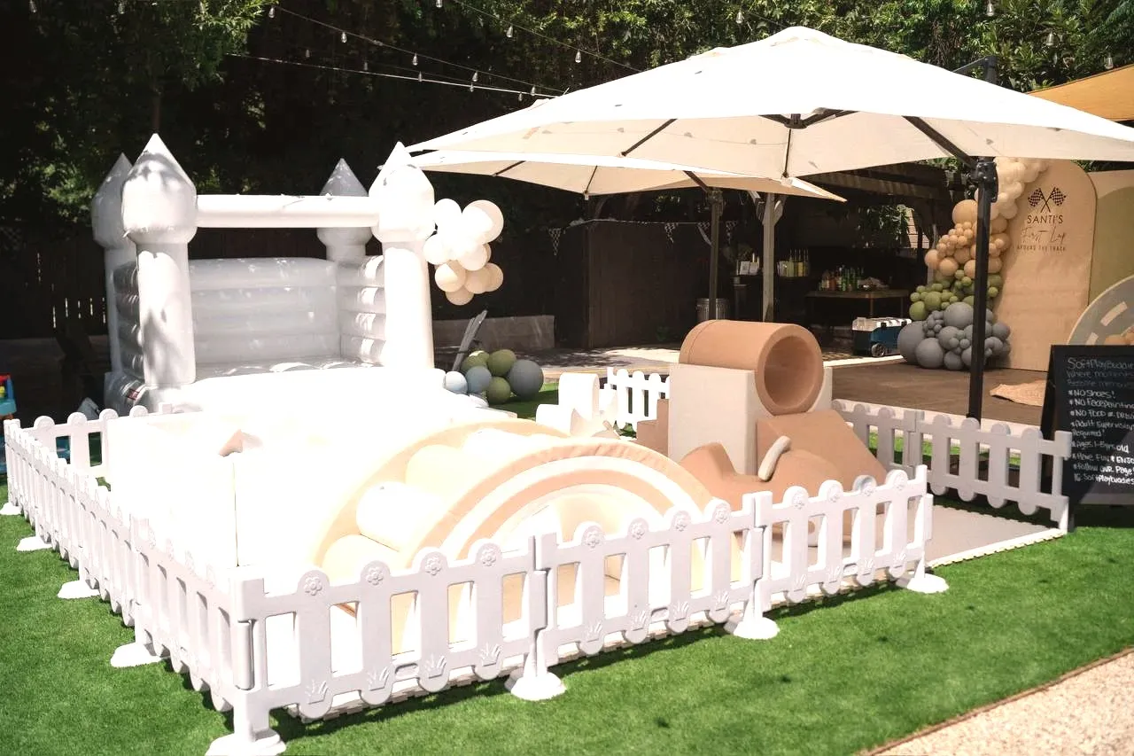 A white bounce house and a soft play set behind a low picket fence on the grass