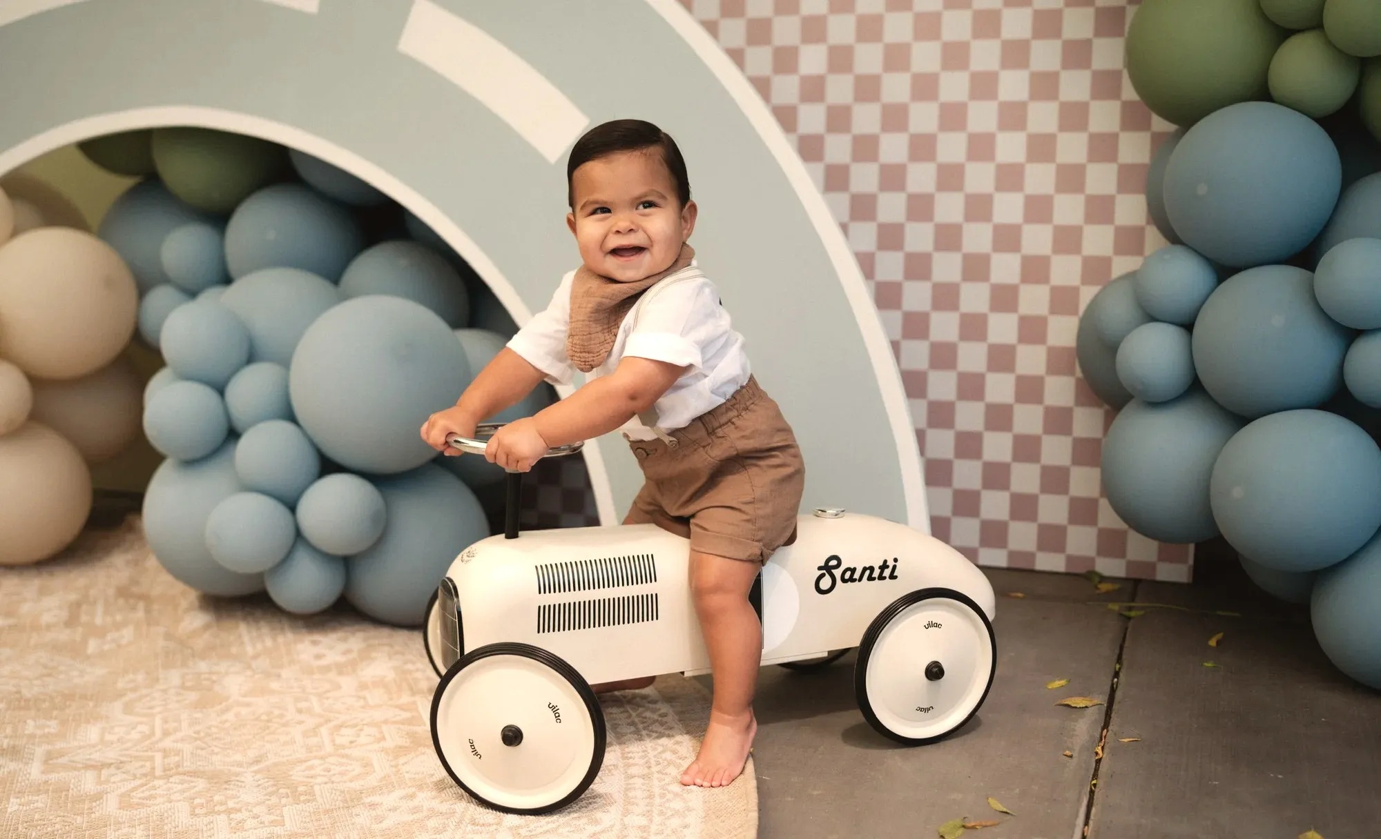 Santi, one year old, grinning on a cream ride-on race car with his name on the side, in front of the race track backdrop