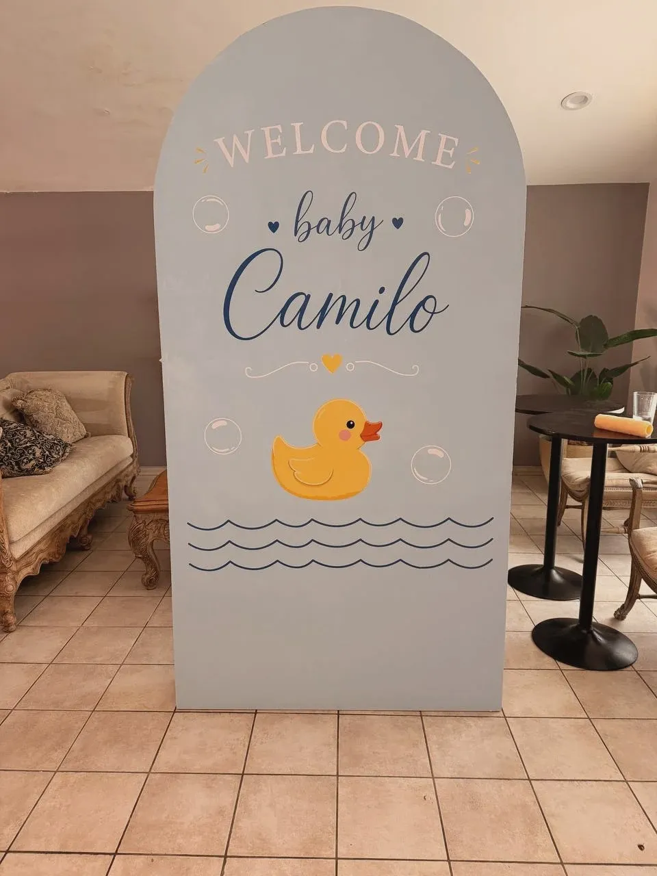 A tall arched wooden backdrop painted pale blue, lettered "Welcome baby Camilo" with a yellow duck and bubbles.