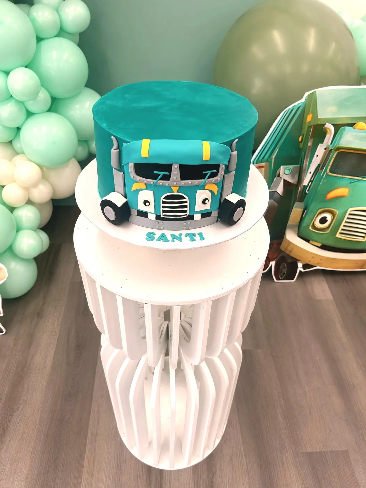 A turquoise fondant cake shaped like the show's trash truck, reading Santi, beside the matching truck cutout