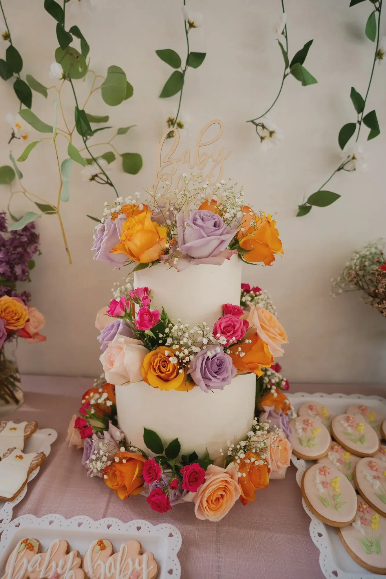 A two-tier white cake ringed with fresh orange, peach, lilac and hot pink roses and baby's breath, in front of a wall of hanging greenery and white blossom