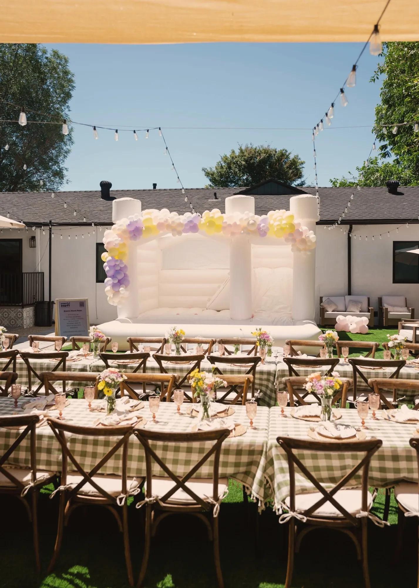 Two long tables in green and cream gingham with wooden cross-back chairs, jars of wildflowers and pink glassware, set facing a white bounce house draped in a lilac and yellow balloon garland