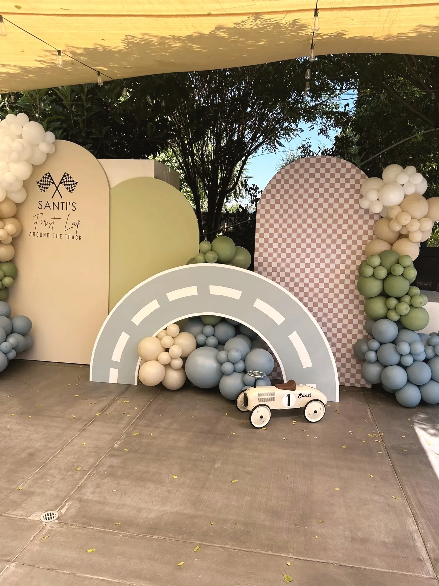 The layered backdrop seen straight on, a Santi's First Lap sign board, a sage arch, a checkered arch, and a curved race track with a cream ride-on race car underneath