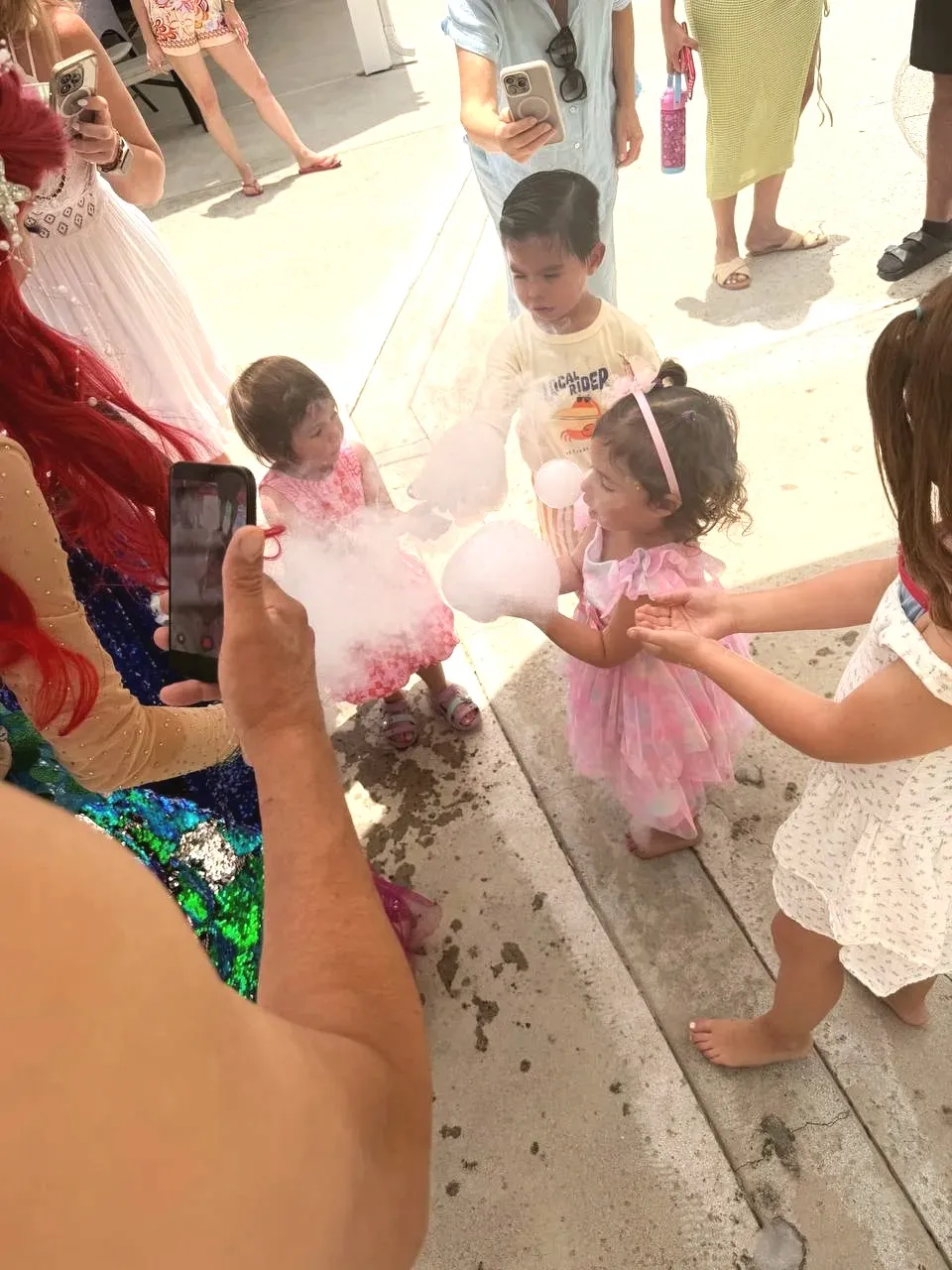 Small children in party dresses standing in a circle on a sunlit patio, each holding a cloud of white foam bubbles in their hands, with the mermaid's sequinned tail at the edge of the frame