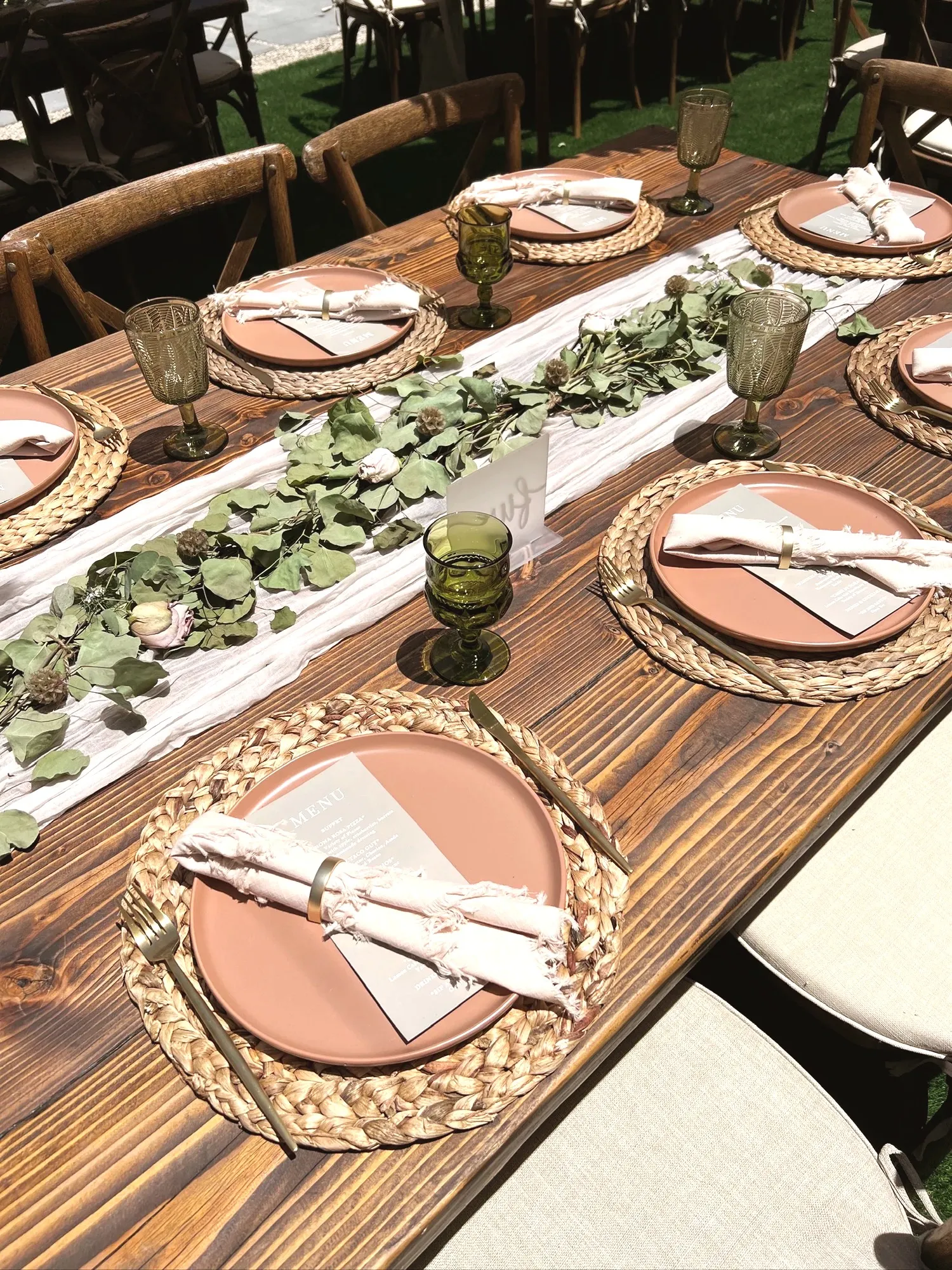 A long farm table set with matching terracotta plates, woven placemats, blush fringed napkins, gold flatware, and green goblets, with a eucalyptus and rose runner down the middle