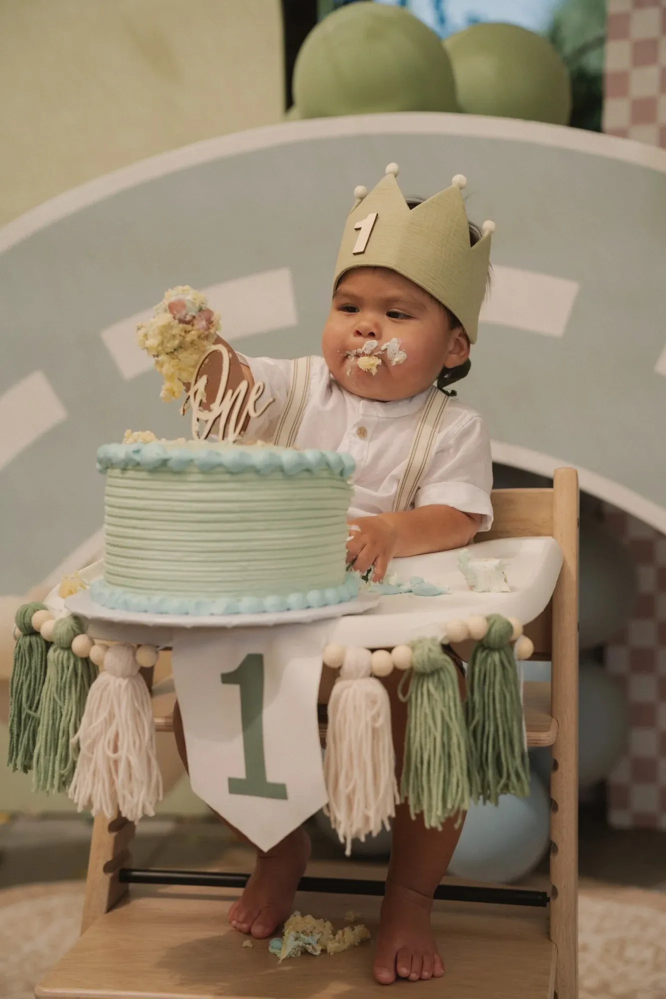 Santi in a green crown in his high chair, digging into a green and blue smash cake