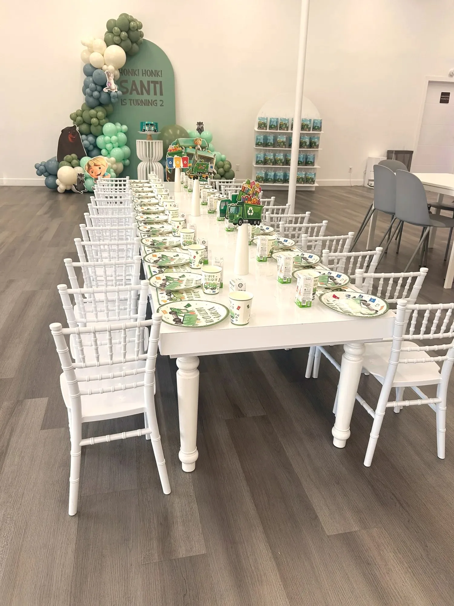 A long white kids' table set with trash-truck plates and lined with little white chairs, the balloon backdrop behind it
