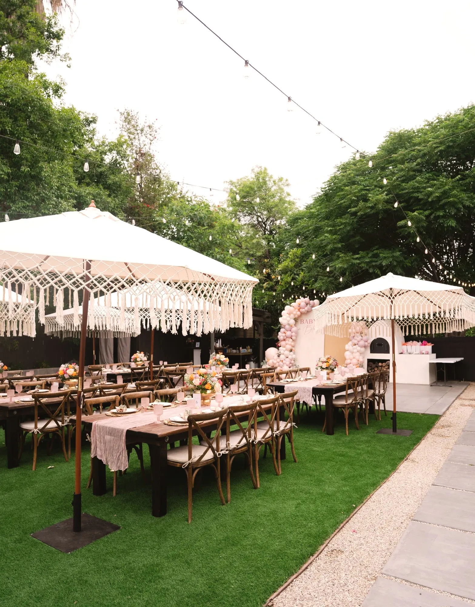 Long wood farm tables on green lawn under cream macramé-fringed umbrellas, set with dusty pink runners and floral centerpieces, with the pink balloon arch at the far end