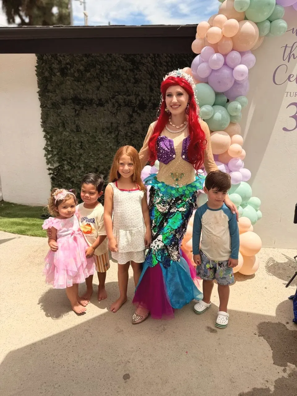 The mermaid standing with four children in front of a green hedge wall and the peach and lavender balloon arch, everyone barefoot on the patio